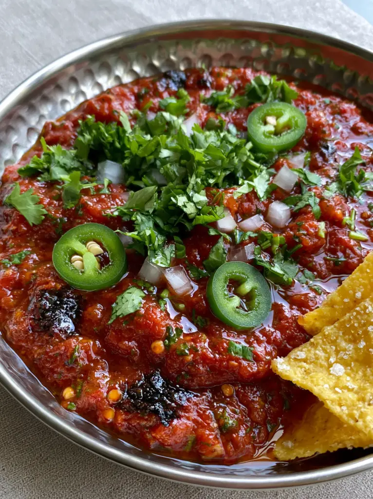 Rustic hammered bowl of Blender salsa sitting on a light gray linen surface with salty tortilla chips.