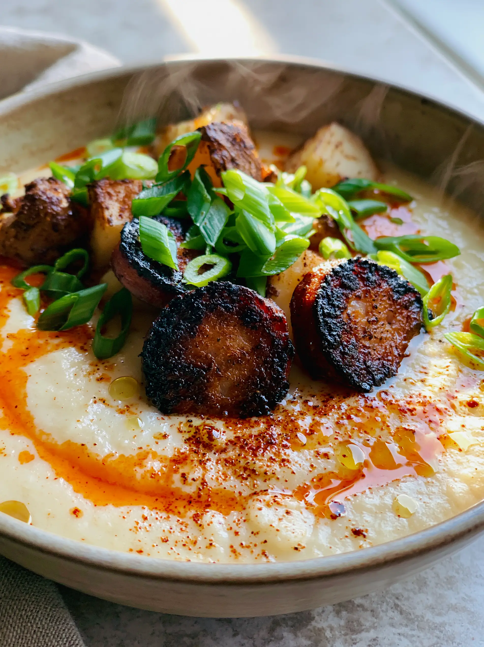 Extreme close-up of velvety ivory cajun potato soup with charred andouille sausage and a vibrant red oil swirl.