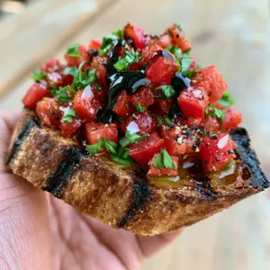 Handheld photo of Tomato Bruschetta showing the deep golden brown sourdough and emerald green basil chiffonade.